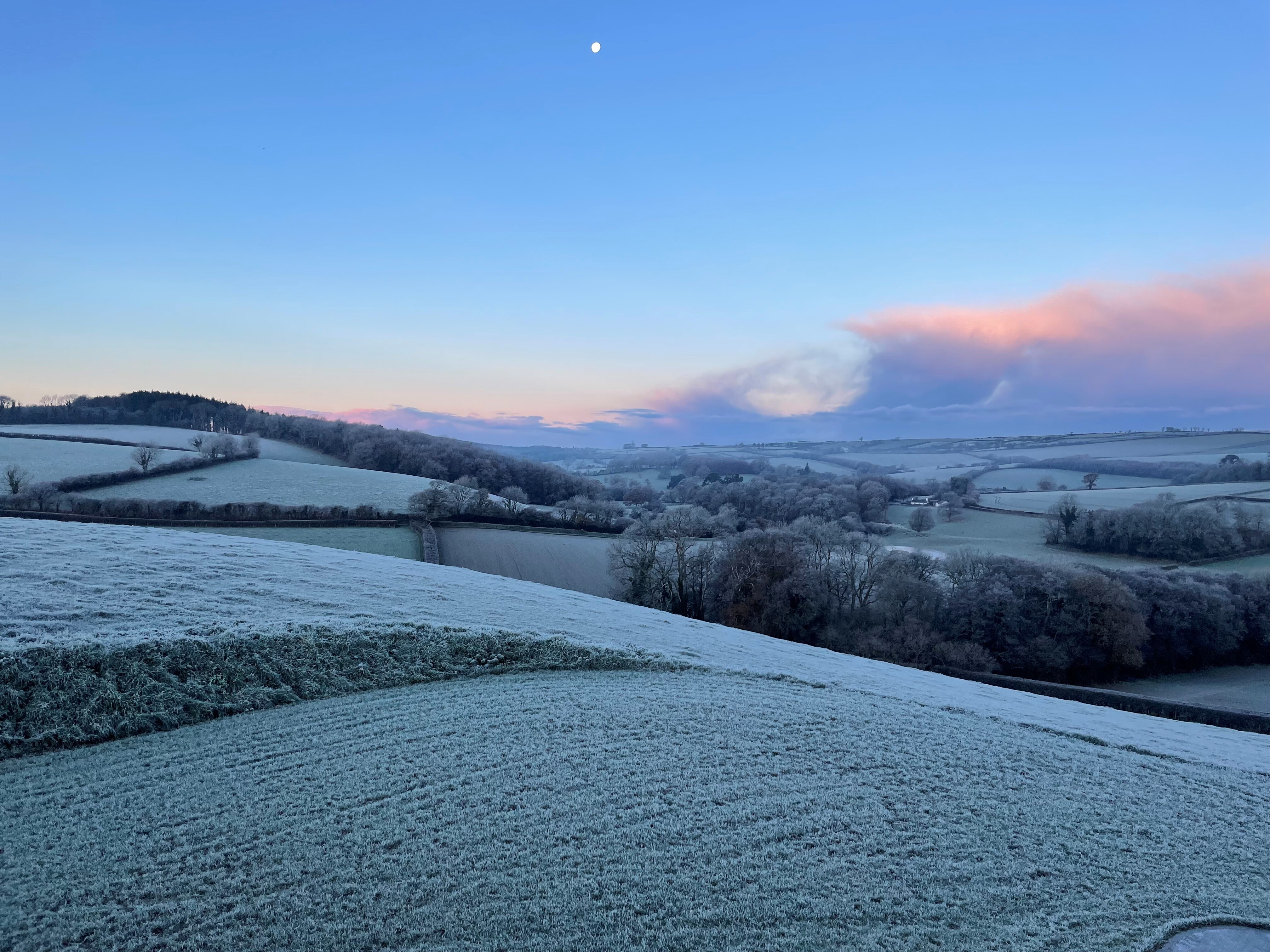 A countryside view from an early morning in winter time, with frost on the fields and the moon still in the sky.