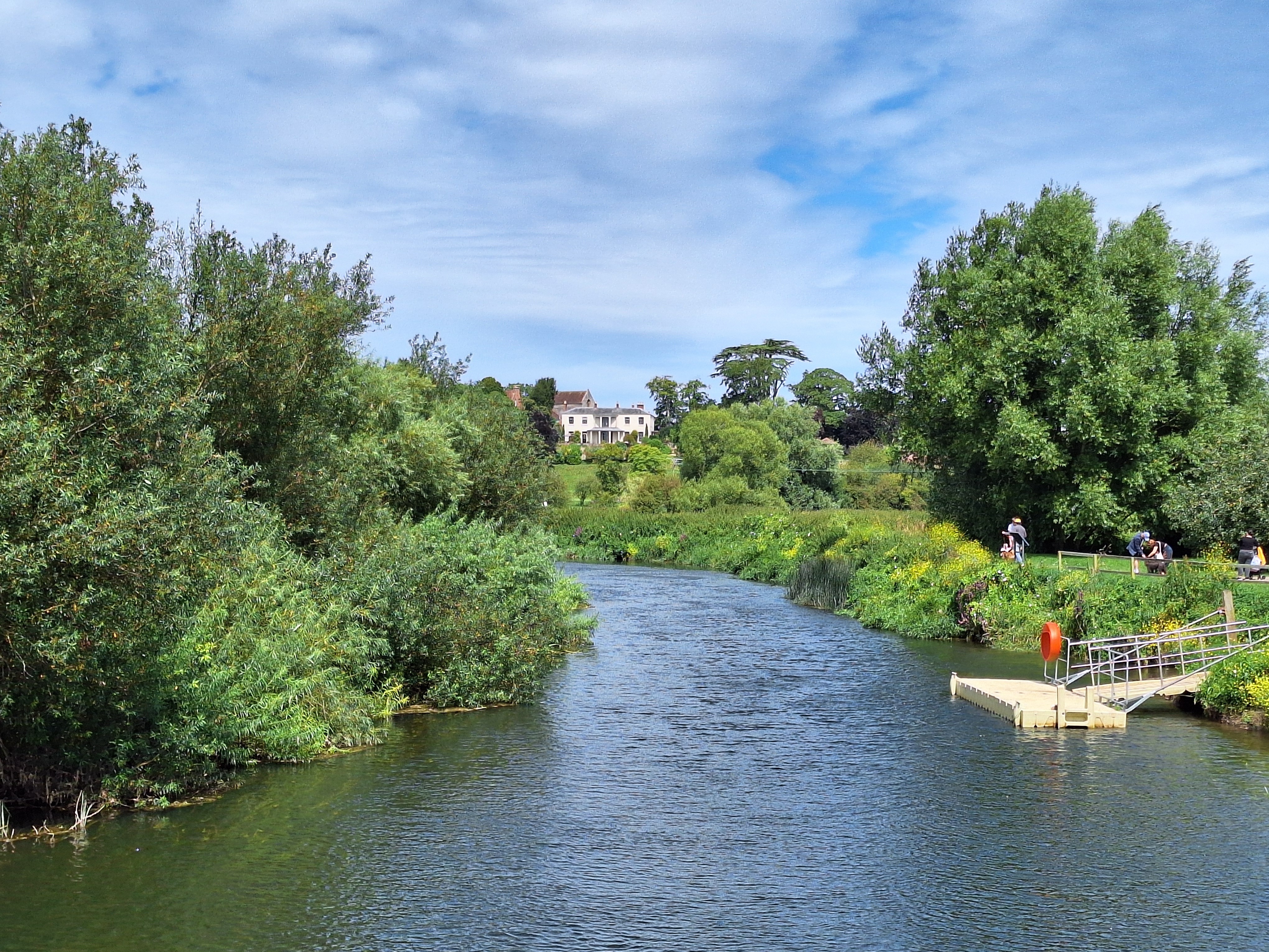 View of a river on a sunny day, surrounded by greenery on each bank along with a small pontoon on one side.