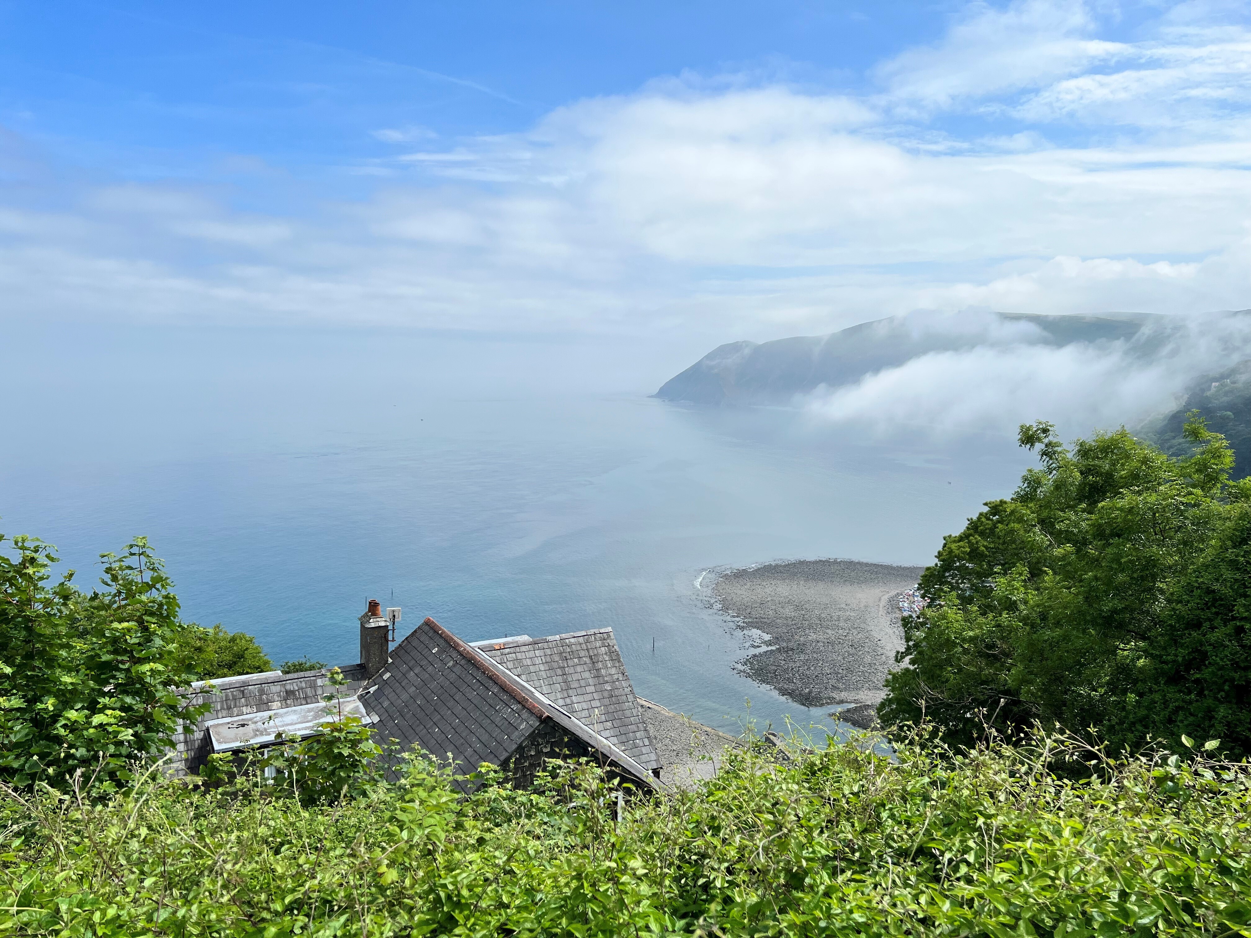 View from the cliff over the top of a house, looking down onto the sea and a pebble beach with a blue sky and soft white clouds.