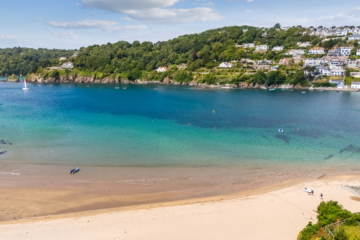 Drone view of the Salcombe Estuary from Mill Bay Beach in East Portlemouth with gorgeous turquoise water and rolling, tree-lined hills.