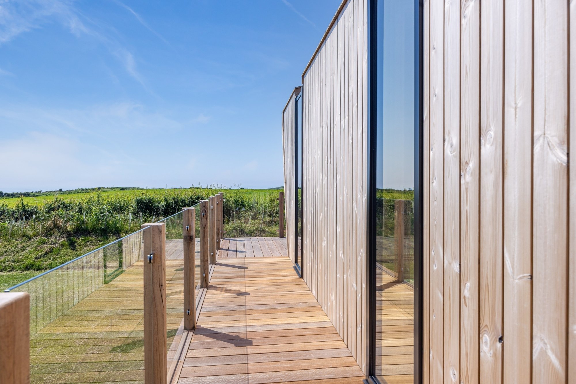 Photo of a wooden, Scandi-style lodge with decking and long windows. Behind is blue skies and a grassy field. 