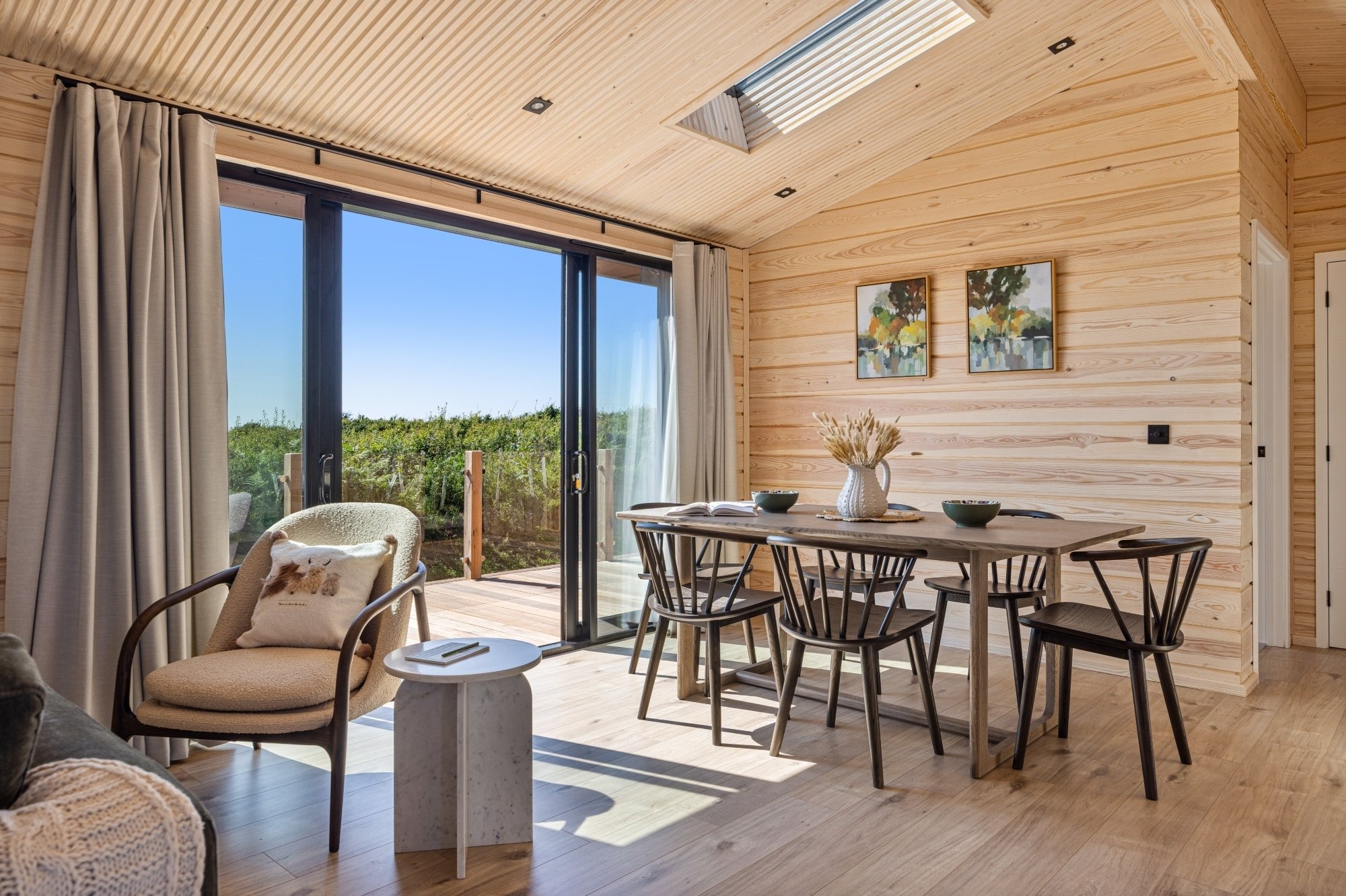 Interior of The Soar - Wooden panelling with balcony doors leading to a terrace and countryside views. Inside is a sun-lit dining table with chairs and an arm chair with side table.